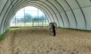 Kobieta z koniem na piaszczystej ujeżdżalni pod łukowatym dachem.