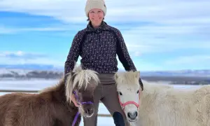Kobieta w zimowej czapce i bluzie stoi między dwoma kucykami na tle zimowego krajobrazu.