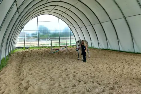 Kobieta z koniem na piaszczystej ujeżdżalni pod łukowatym dachem.