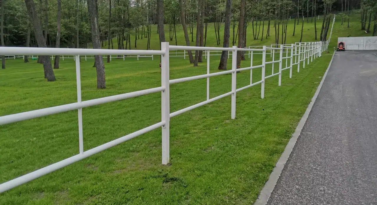 Biały, metalowy padok otoczony zieloną trawą i drzewami. W tle widać drogę i pomarańczowy traktor.
